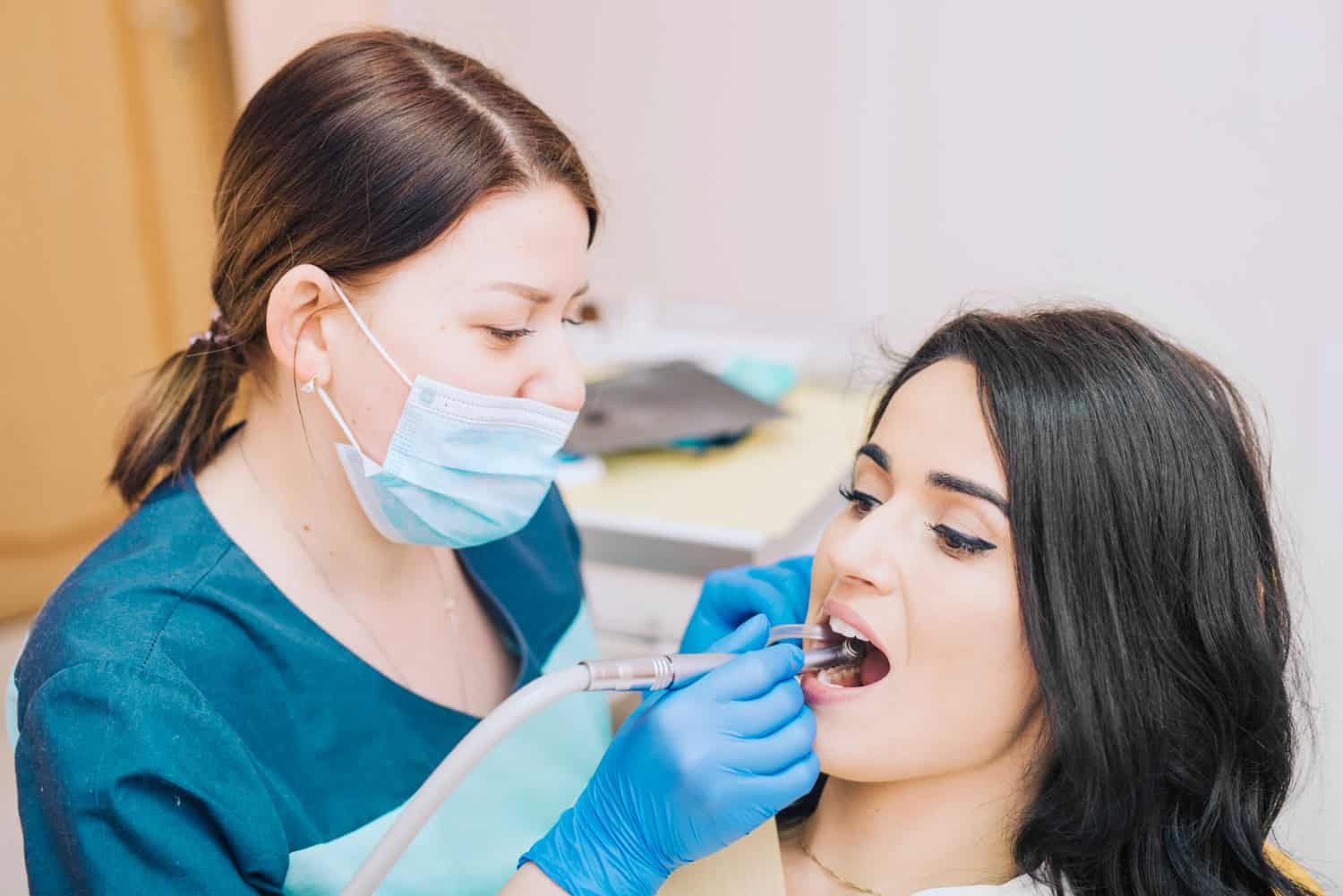 Dentist wearing gloves and a face mask performing a dental examination on a female patient using a dental handpiece in a modern dental clinic.